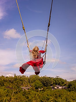 Bali swing trend. Caucasian woman in long red dress swinging in the jungle rainforest. Vacation in Asia. Travel lifestyle. Blue