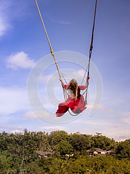 Bali swing trend. Caucasian woman in long red dress swinging in the jungle rainforest. Vacation in Asia. Travel lifestyle. Blue
