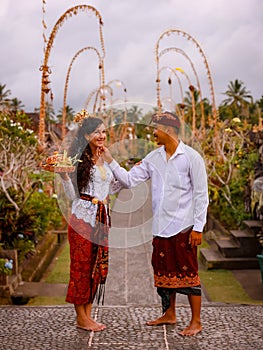 Balinese ceremony. Multicultural couple going to Hindu religious ceremony with god`s offerings. Penjor bamboo decoration.