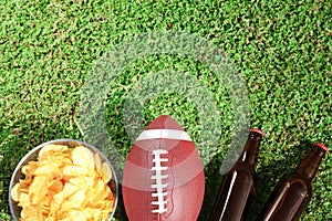 Ball for American football, beverage and chips on fresh green field grass, flat lay.