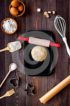 Ball of fresh raw dough near ingedients and cookware on dark wooden background top view