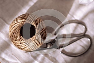 A ball of natural brown jute twine for needlework,or household needs lies next to vintage scissors on a white fabric.
