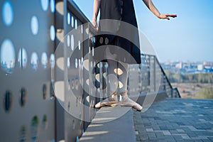 Ballerina in ballet legs in shoes and black tutu dancing by the fence. Beautiful young woman in black dress and pointe dancing