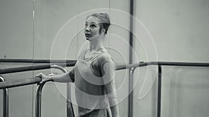 Ballerina in beige sweatsuit and pointe stretches on barre in ballet gym. Woman standing near bar and mirror, preparing