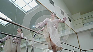 Ballerina in beige sweatsuit and pointe stretches on barre in ballet gym. Woman standing near bar and mirror, preparing