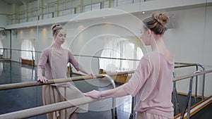 Ballerina in beige sweatsuit and pointe stretches on barre in ballet gym. Woman standing near bar and mirror, preparing