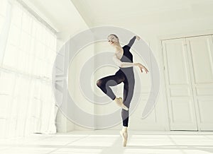 ballerina in a black bodysuit and total black tights in a room by the window shows the elements and steps of ballet