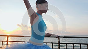 Ballerina in blue ballet tutu and point on stone quay above ocean or sea at sunrise. Young beautiful brunette woman
