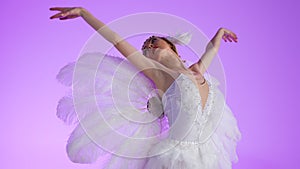 A ballerina in a chic image of a white swan is gracefully dancing on a purple background in a light studio. Festive