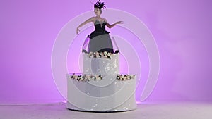 Ballerina dressed as a black swan emerges from a shiny white cake. Young women in white tutus are dancing around