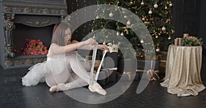 Ballerina ties up pointes sitting in a Christmas decorated room