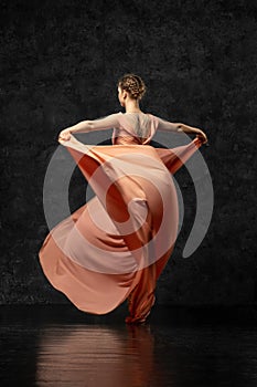 Ballerina turned her back and dancing in a long peach dress on a black background.