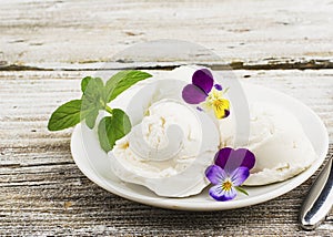 Balls of creamy vanilla home ice cream on a ceramic white saucer with edible flowers on a wooden light background