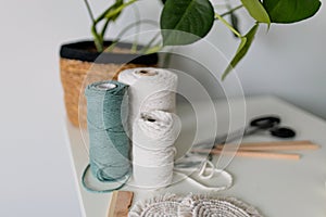 Balls of white and green yarn with pot of monstera on white table. Handmade macrame braiding. Female hobby. Selective