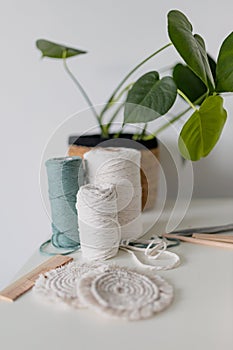 Balls of white and green yarn with pot of monstera on white table. Handmade macrame braiding. Female hobby. Selective