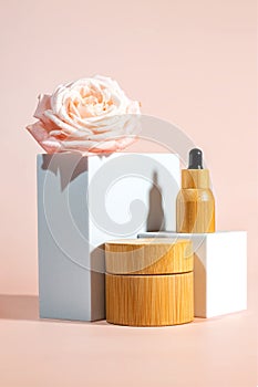 Bamboo cosmetic bottles with pipette and cream jar on white podium, product packaging with rose flower, anti aging serum