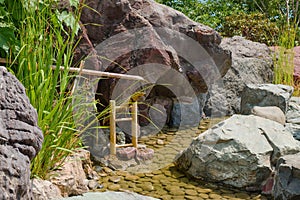 Bamboo sound water fountain next to calm languid stream surrounded stones as part of traditional Japanese Zen Garden