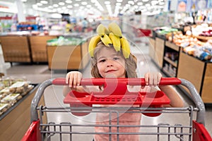 Banana on funny child head. Funny kid with shopping cart buying food at grocery store or supermarket. Surprised kids