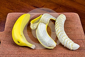 Banana, peeled banana and sliced banana on a wooden cutting board in front view