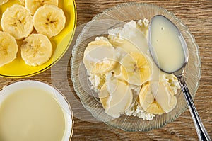 Banana in saucer, bowl with condensed milk, spoon in bowl with cottage cheese, slices of banana and condensed milk on table. Top