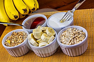 Banana sliced on ramekin and fork dipping a banana slice into yogurt with oatmeal, granola, plain yogurt and honey as side dishes