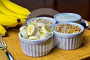 Banana sliced ramekin with oatmeal, granola and plain yogurt as side dishes under bamboo mat with bunch of bananas