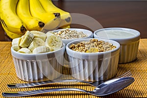 Banana sliced ramekin with oatmeal, granola and plain yogurt as side dishes under bamboo mat with bunch of bananas