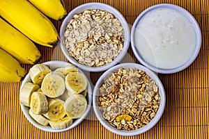 Banana sliced ramekin with oatmeal, granola and plain yogurt as side dishes under bamboo mat with bunch of bananas besides