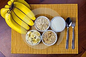 Banana sliced ramekin with oatmeal, granola and plain yogurt as side dishes under bamboo mat with bunch of bananas besides