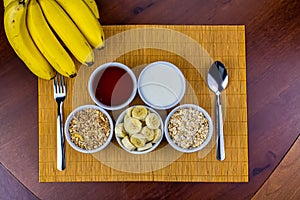 Banana sliced ramekin with oatmeal, granola, plain yogurt and honey as side dishes under bamboo mat with bunch of bananas