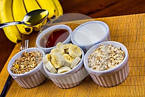 Banana sliced ramekin with oatmeal, granola, plain yogurt and honey as side dishes under bamboo mat with bunch of bananas