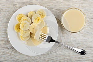 Banana strung on fork, white plate with slices of banana and condensed milk, bowl with milk on table. Top view