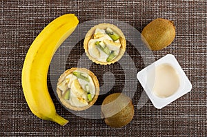 Banana, tartlets with kiwi, banana and yogurt, kiwi fruits, jar with yogurt on mat. Top view