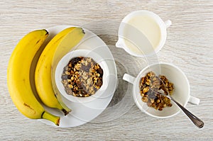 Bananas, bowl with muesli in plate, pitcher with yogurt, spoon in bowl with granola on wooden table. Top view