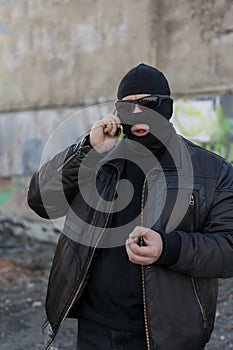 A bandit in a black leather jacket and a mask talking on the phone on the street near an abandoned building