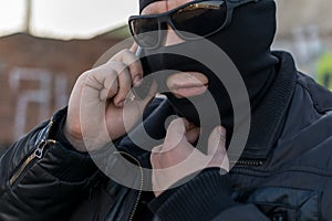 A bandit in a black leather jacket and a mask talking on the phone on the street near an abandoned building