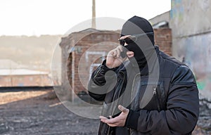 A bandit in a black leather jacket and a mask talking on the phone on the street near an abandoned building