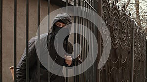 Bandit man in black mask and hood with baseball bat crawls through fence mesh