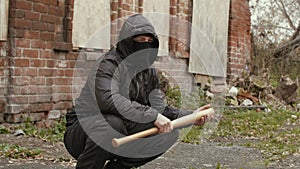 Bandit man in black mask and jacket with hood with baseball bat sits on street
