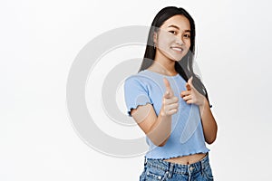 Bang. Smiling asian girl pointing finger pistols at camera, choosing you, recruiting people, inviting to an event