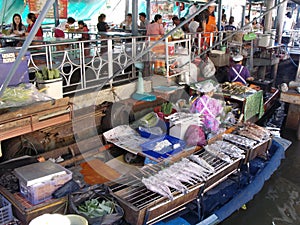 People eating at tables the fish that a woman cooks in her boat at the Taling Chan Floating Market in Bangkok