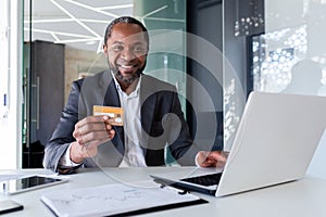 Bank employee, smiling young African American man sitting in office at desk with laptop, holding and showing credit card