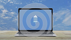 Bank icon on modern laptop computer monitor screen on wooden table over blue sky with white clouds, Business banking online c