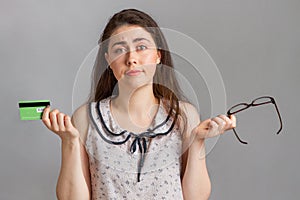 Bank investments and cards. A young woman in a blouse, holding a Bank card and glasses, in bewilderment spreading her hands apart