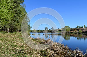 The bank of the river on a summer day. Blue sky and blue calm water. Trees are reflected in water