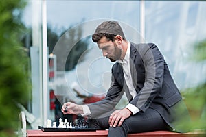 Banker male businessman playing chess sitting on bench near office, pensive making strategic move