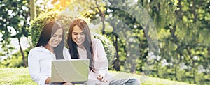 Banner Asian two women talking using laptop sit on green meadow at green park. Panorama Friendship girls chat, talk look at laptop