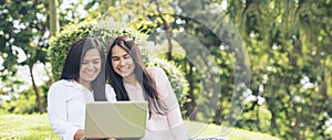 Banner Asian two women talking using laptop sit on green meadow at green park. Panorama Friendship girls chat, talk look at laptop