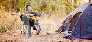 Banner format image of smiling young man in black hat sitting near touristic tent and playing guitar in forest
