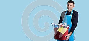 Banner, long format on blue background. Young serious caucasian man carrying a bucket with diverse cleaning products